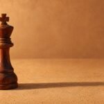 A single hand-carved wooden chess king standing on a plain sand-coloured surface, warm directional light from the left, a soft long shadow, and.