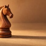 A hand-carved wooden chess knight standing on a plain sand-coloured surface, warm directional light from the left, a soft long shadow, and plenty of.