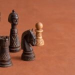 A warm natural-light photograph of an African-designed wooden chess set on a plain warm background, with a few pieces arranged to suggest a beginner.