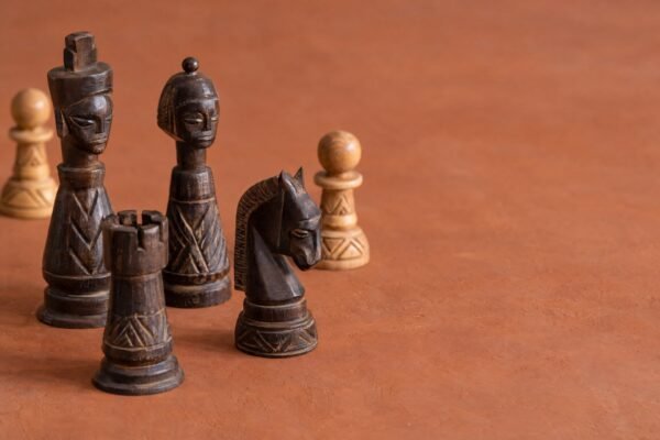 A warm natural-light photograph of an African-designed wooden chess set on a plain warm background, with a few pieces arranged to suggest a beginner.