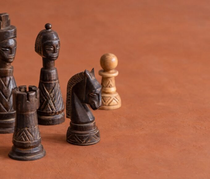 A warm natural-light photograph of an African-designed wooden chess set on a plain warm background, with a few pieces arranged to suggest a beginner.