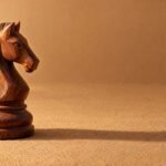 A hand-carved wooden chess knight standing on a plain sand-coloured surface, warm directional light from the left, a soft long shadow and plenty of.