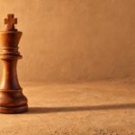 A hand-carved wooden chess king standing alone on a plain sand-coloured surface, warm directional light from the left, a soft long shadow and.