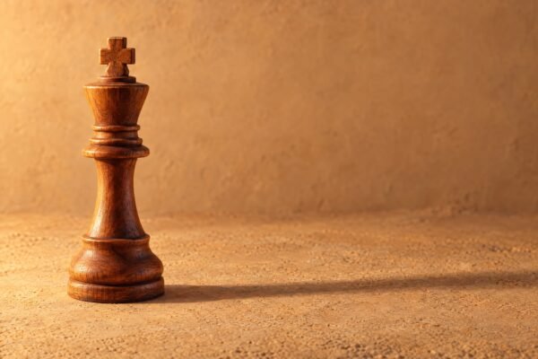 A hand-carved wooden chess king standing alone on a plain sand-coloured surface, warm directional light from the left, a soft long shadow and.