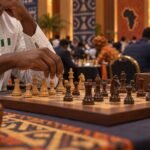 Odion Aikhoje’s hands poised over a wooden chessboard during a tense board-two game in a crowded Olympiad hall, with out-of-focus players and Adire.