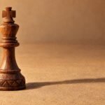 A hand-carved wooden chess king standing alone on a plain sand-coloured surface, warm directional light from the left, a soft long shadow, and plenty.