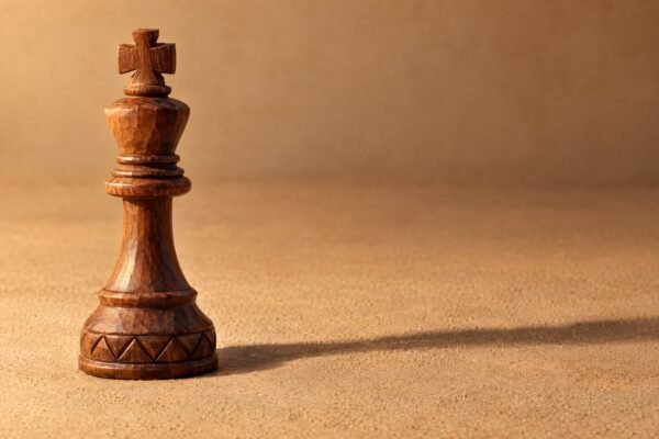 A hand-carved wooden chess king standing alone on a plain sand-coloured surface, warm directional light from the left, a soft long shadow, and plenty.