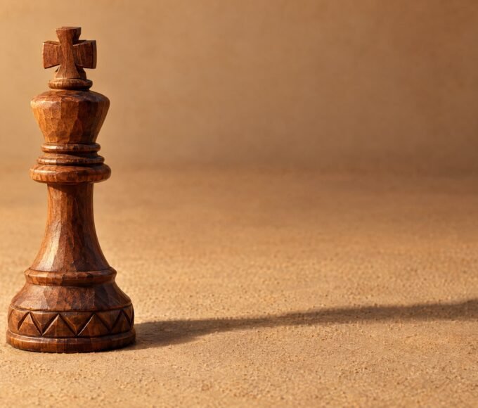 A hand-carved wooden chess king standing alone on a plain sand-coloured surface, warm directional light from the left, a soft long shadow, and plenty.