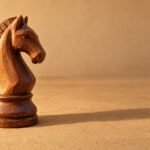 A hand-carved wooden chess knight standing on a plain sand-coloured surface, warm directional light from the left, a soft long shadow, and plenty of.