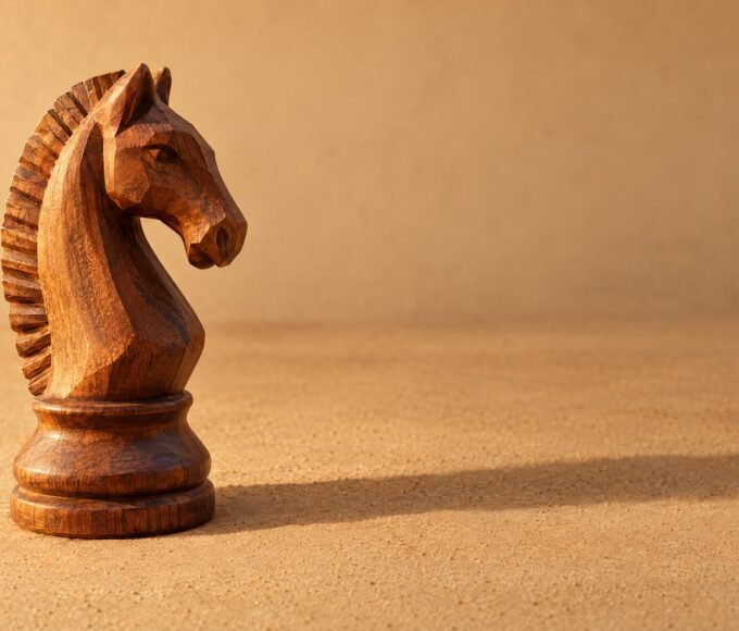 A hand-carved wooden chess knight standing on a plain sand-coloured surface, warm directional light from the left, a soft long shadow, and plenty of.