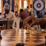 A chess player’s hand carefully guides a king beside a lone passed pawn on a wooden board in a busy Nigerian chess hall, with out-of-focus players.