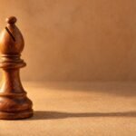 A hand-carved wooden chess bishop standing on a plain sand-coloured surface, warm directional light from the left, a soft long shadow, and plenty of.