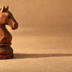 A hand-carved wooden chess knight standing on a plain sand-coloured surface, warm directional light from the left, a soft long shadow and plenty of.