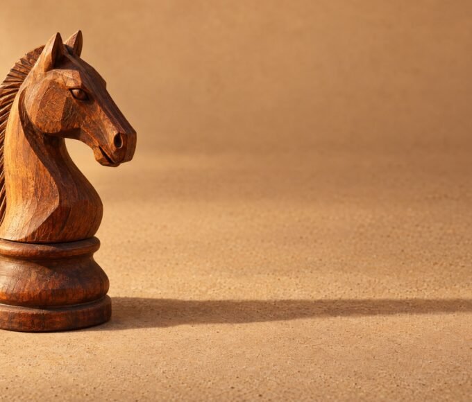 A hand-carved wooden chess knight standing on a plain sand-coloured surface, warm directional light from the left, a soft long shadow and plenty of.
