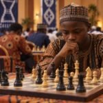 A young Nigerian chess player studies a tense middlegame at board level in a busy tournament hall, with the subject sharp in the foreground,.