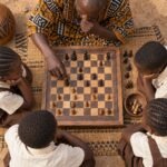 An overhead flat-lay photograph of a primary-school chess lesson in Africa, with a wooden board and hands considering a position, surrounded by.