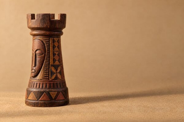 A hand-carved wooden chess rook decorated with Nok-inspired geometric carving, standing on a warm sand-coloured surface